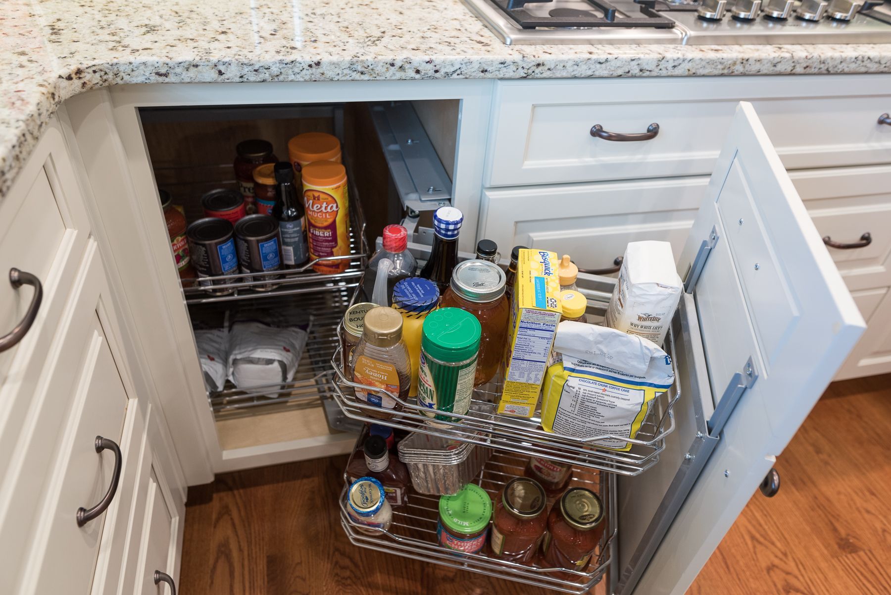 Pull-out kitchen cabinet with stainless steel shelves holding spices, jars, and snacks. White cabinets, countertop.