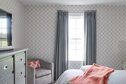 Bedroom with gray dresser, chair, window, patterned wallpaper, and coral blanket.