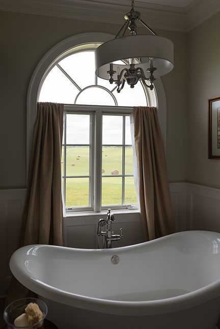 A white bathtub in front of a window with tan curtains, a chandelier hangs overhead.