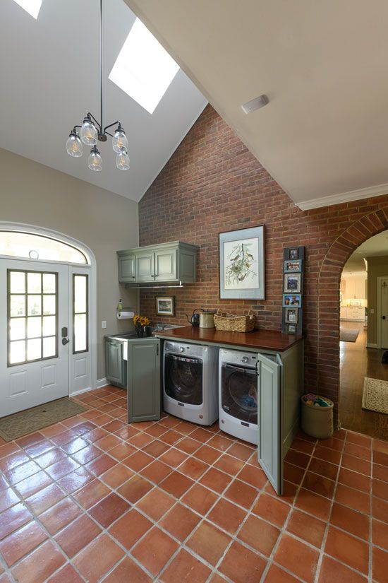 Laundry room with red brick wall, terracotta tiles, white washer/dryer, and green cabinets. Skylight brightens the space.
