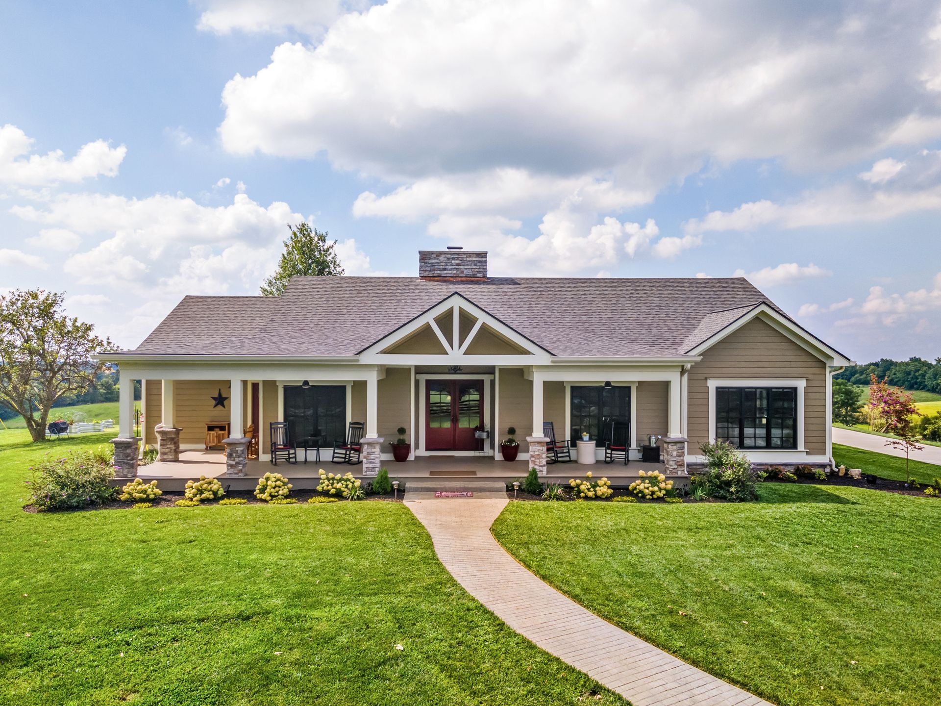 Tan farmhouse with wrap-around porch, red front door, brick walkway, green lawn, and cloudy sky.
