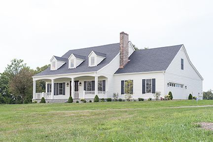 White house with porch, black shutters, three dormers, and a brick chimney on a grassy lawn.