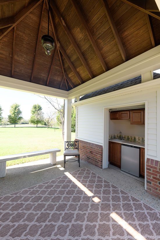 Covered outdoor patio with rug, seating, and small kitchen overlooking a grassy field.