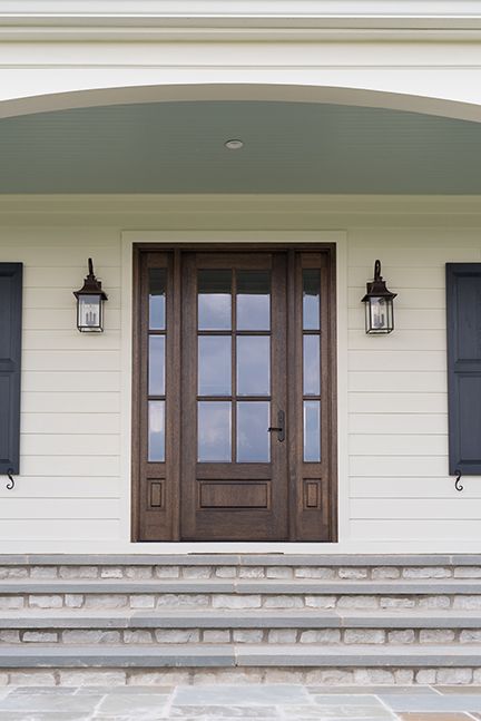 Wooden front door with sidelights, surrounded by cream-colored siding, flanked by sconces and black shutters. Stone steps lead up.