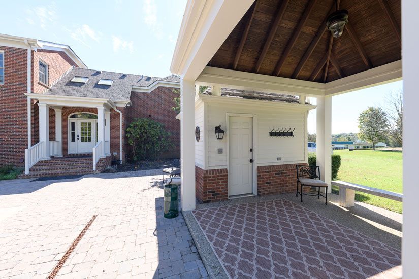 Brick building with a covered patio. A door and bench are beneath the patio's wood-paneled roof, overlooking a grassy field.