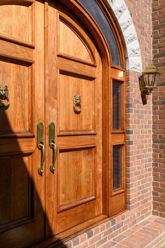Wooden double doors with arched window and sidelight, gold hardware, red brick wall.