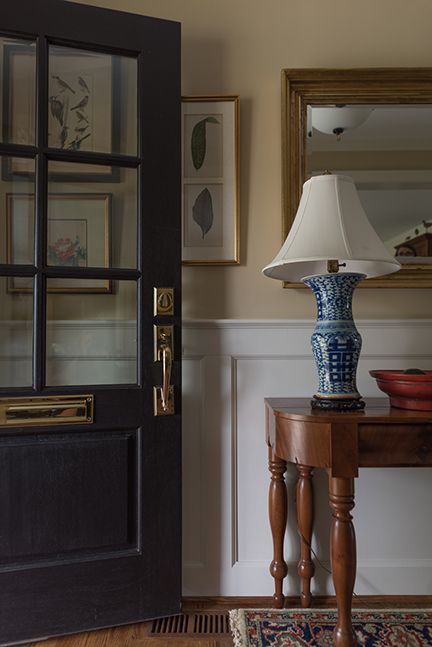 Black door with glass panels opens to a hallway with a table, lamp, mirror, and framed art.