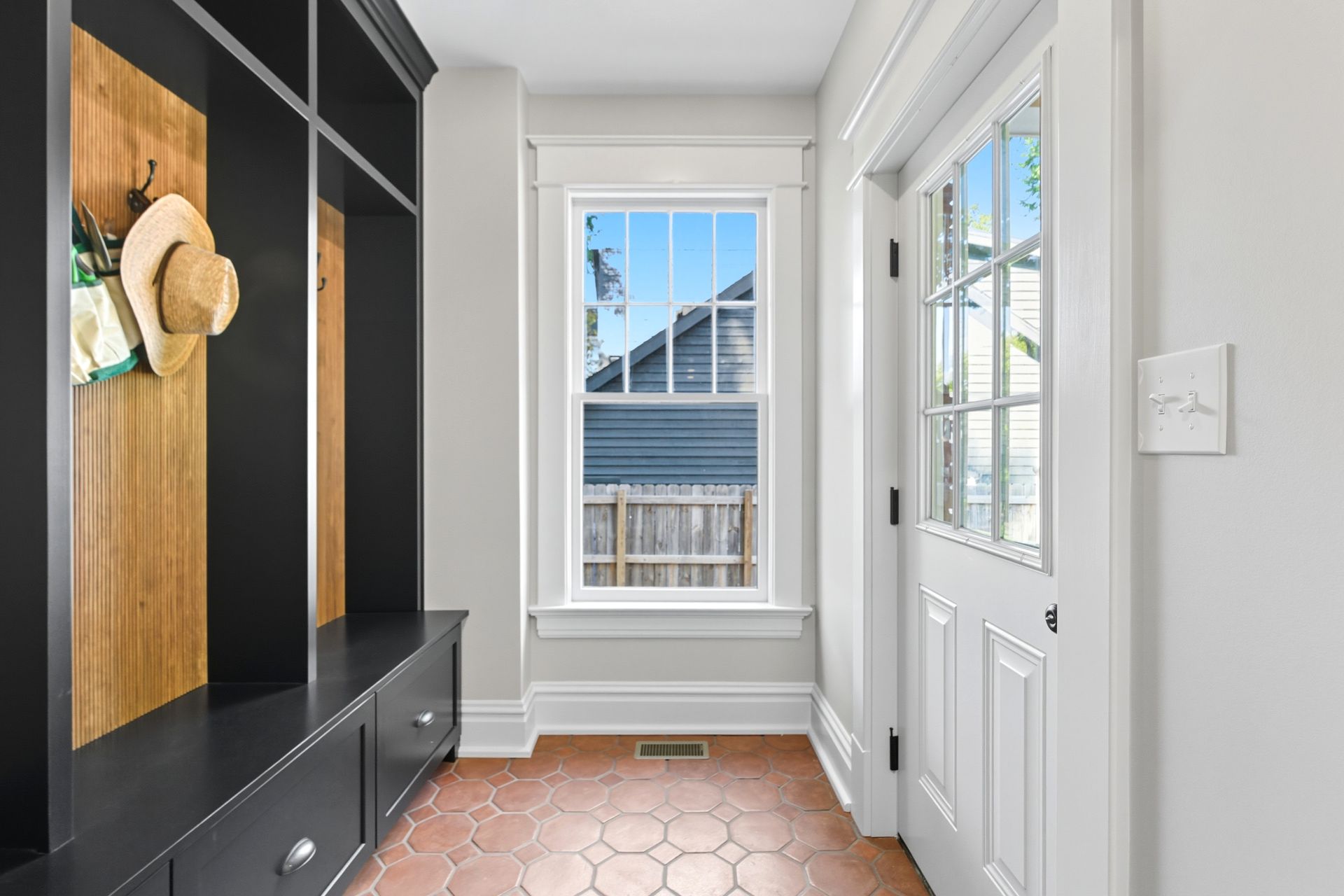 Entryway with black built-in bench and cabinets, terracotta tile floor, white door, and small window showing exterior.