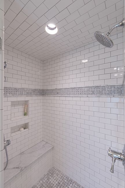 White tiled shower with built-in bench, mosaic tile stripe, recessed shelf, and rain shower head.