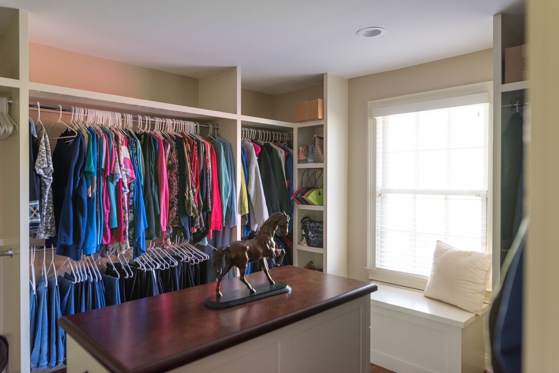 Walk-in closet with clothing racks, wooden island, and window seat with pillow and horse statue.