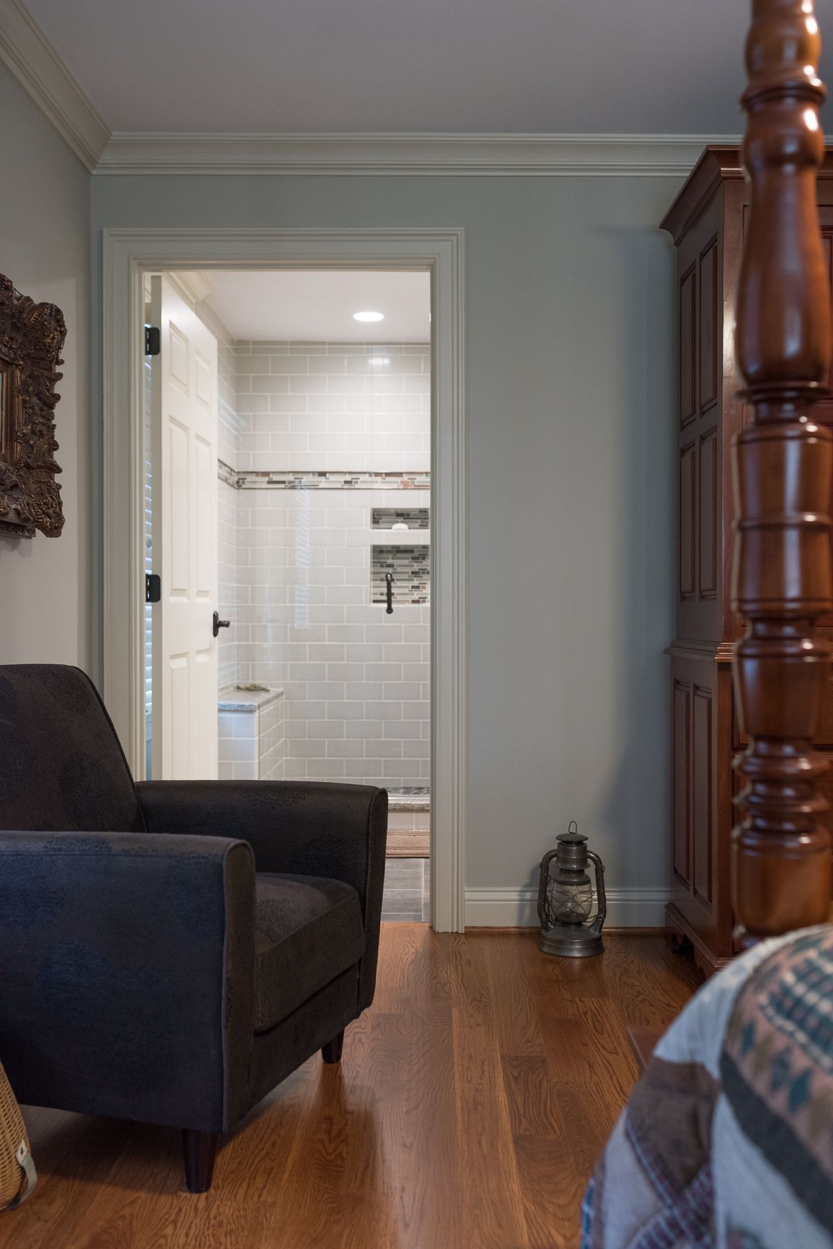 Bedroom with a dark armchair, open doorway to a bathroom with white tiles, and a wooden armoire.
