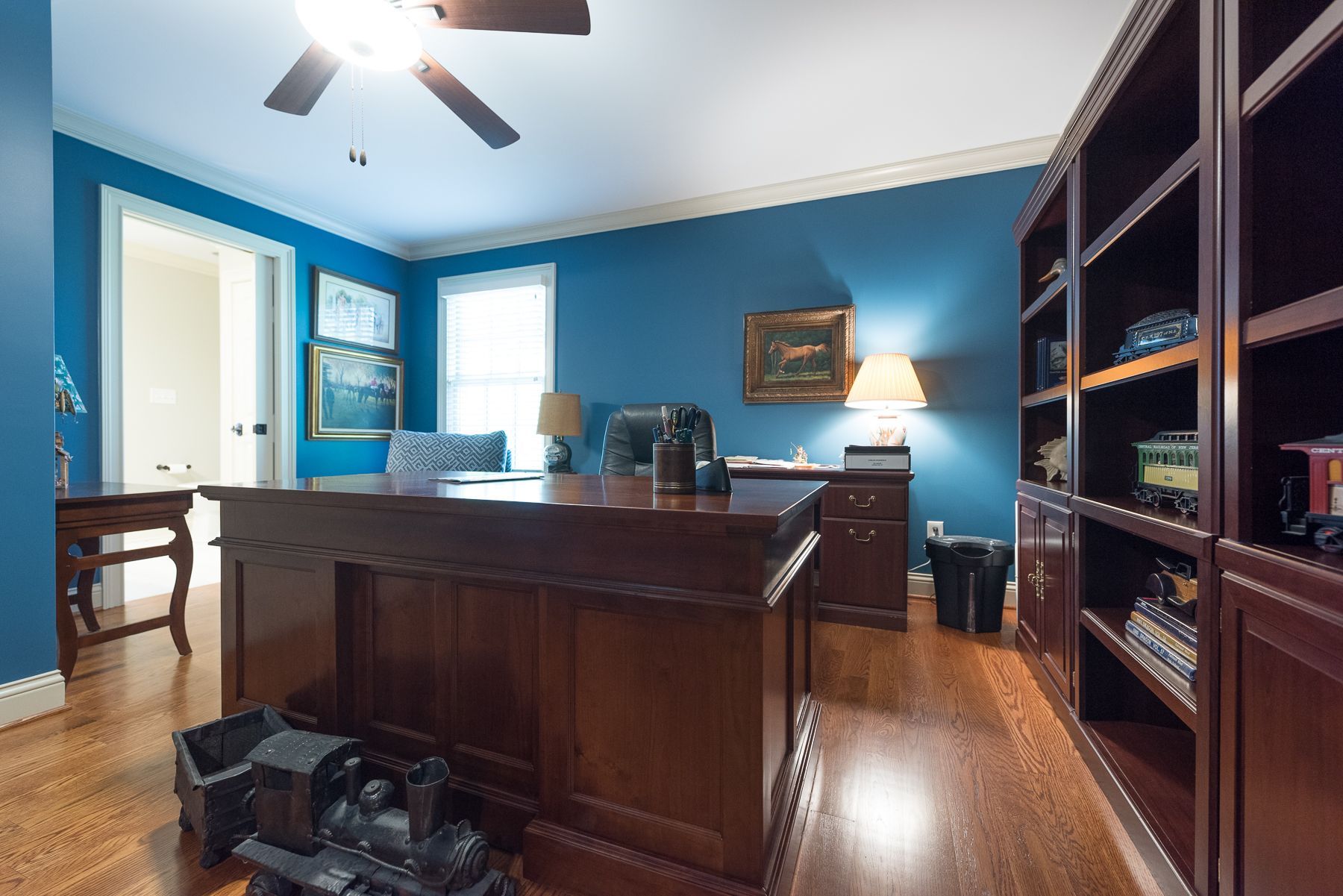 Home office with blue walls, brown desk, wooden bookshelves, and train set on floor.