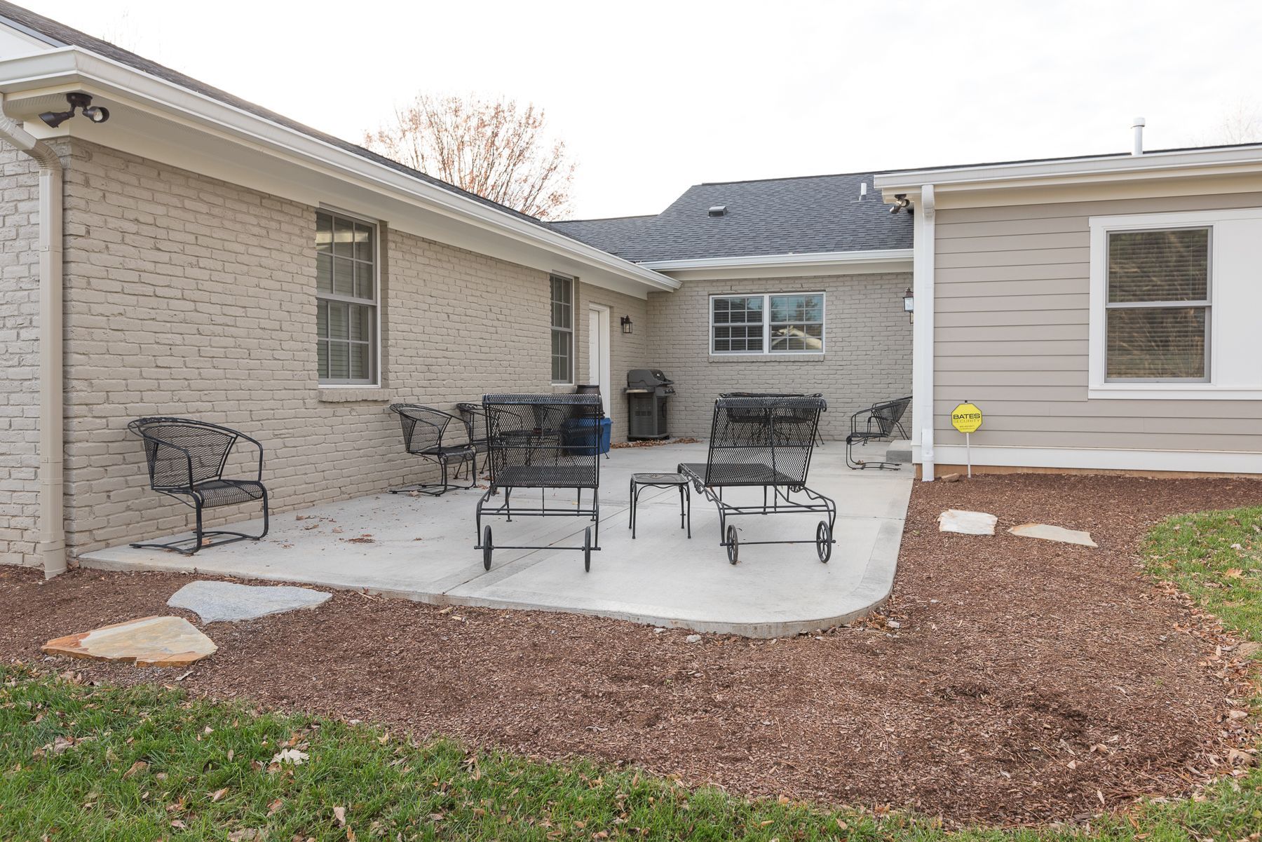 Patio with black metal furniture, two brick buildings, and a gray roof. Brown mulch and green grass surround.