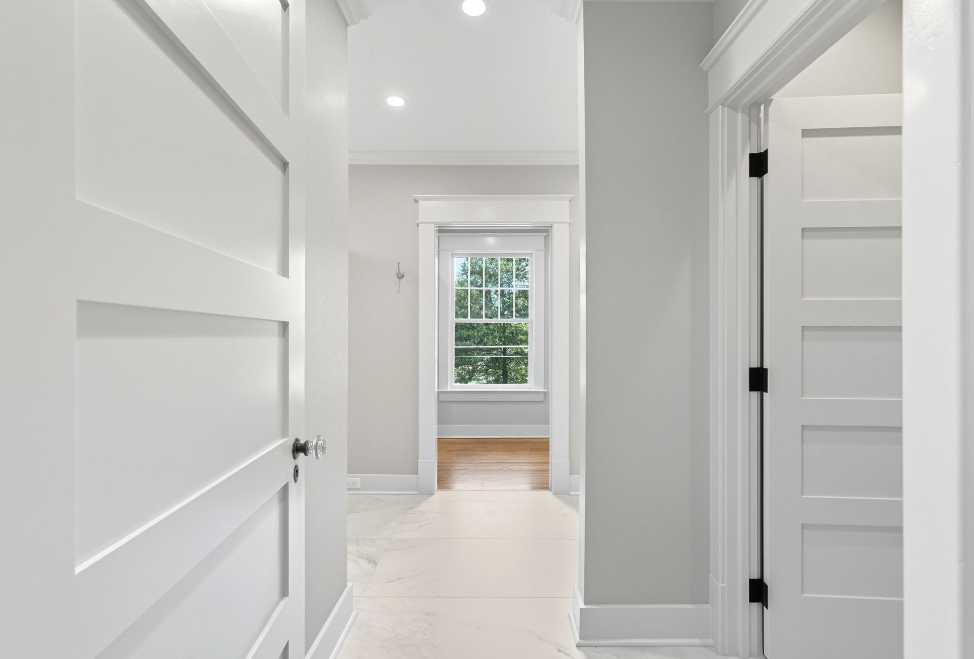 Bright hallway with white doors, gray walls, a window, and light wood floors.