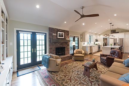 Spacious living room with a brick fireplace, open to a kitchen. Includes seating, rug, and a ceiling fan.