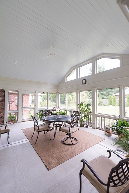 Enclosed sunroom with a table and chairs, windows, plants, and a rug. White walls and ceiling.