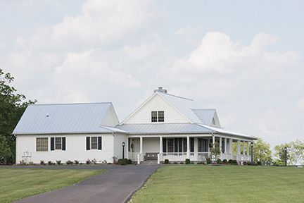 White farmhouse with a porch and gray roof on a green lawn, black driveway, and a cloudy sky.