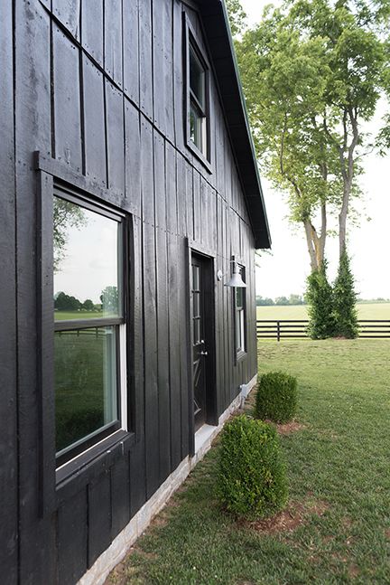 Black exterior of a building with white-trimmed windows and door; green lawn with trees and fence in the background.