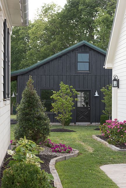Exterior view of a black barn-like structure with a green roof, framed by white houses and garden beds.
