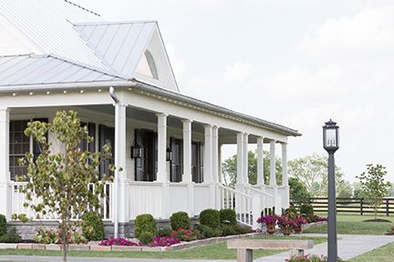 White farmhouse with a wraparound porch, lush landscaping, and a black lamp post against a cloudy sky.