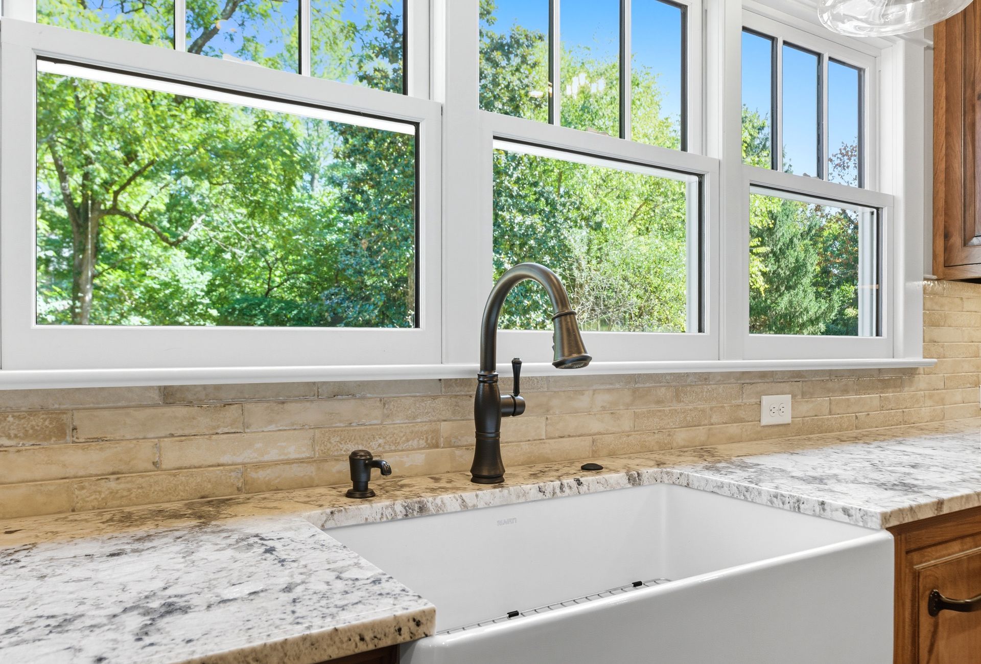 White farmhouse sink with dark faucet and granite countertop under window overlooking trees.