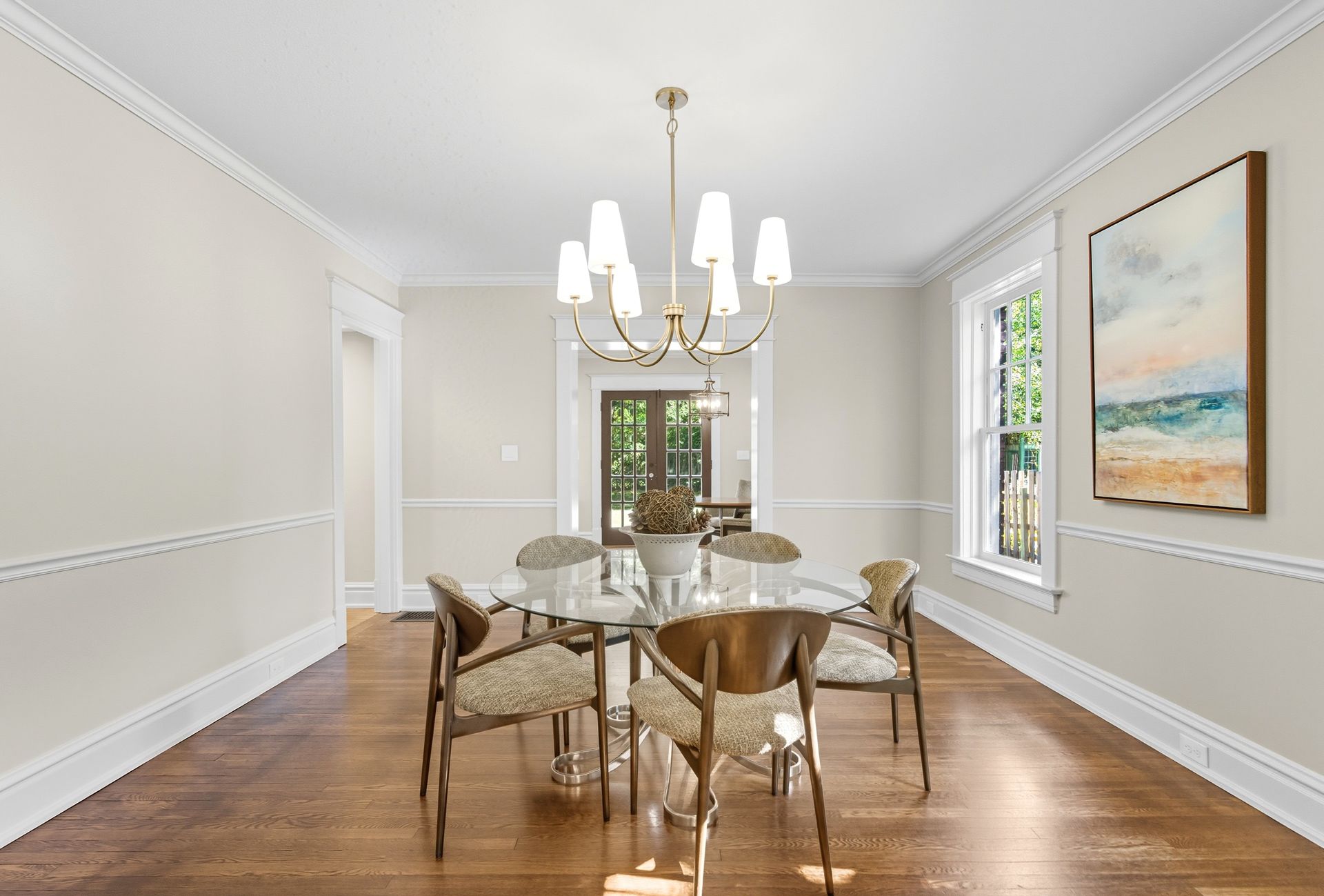 Dining room with a glass table, chairs, and artwork; bright and airy with a chandelier.