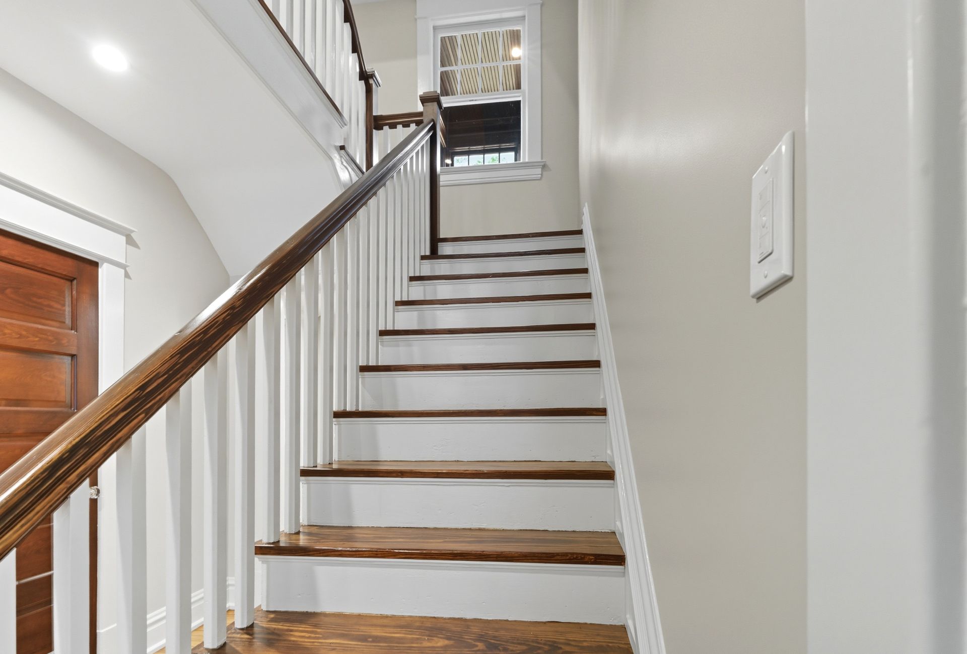 Staircase with wooden steps, white risers, and brown handrail, leading upwards toward a window.