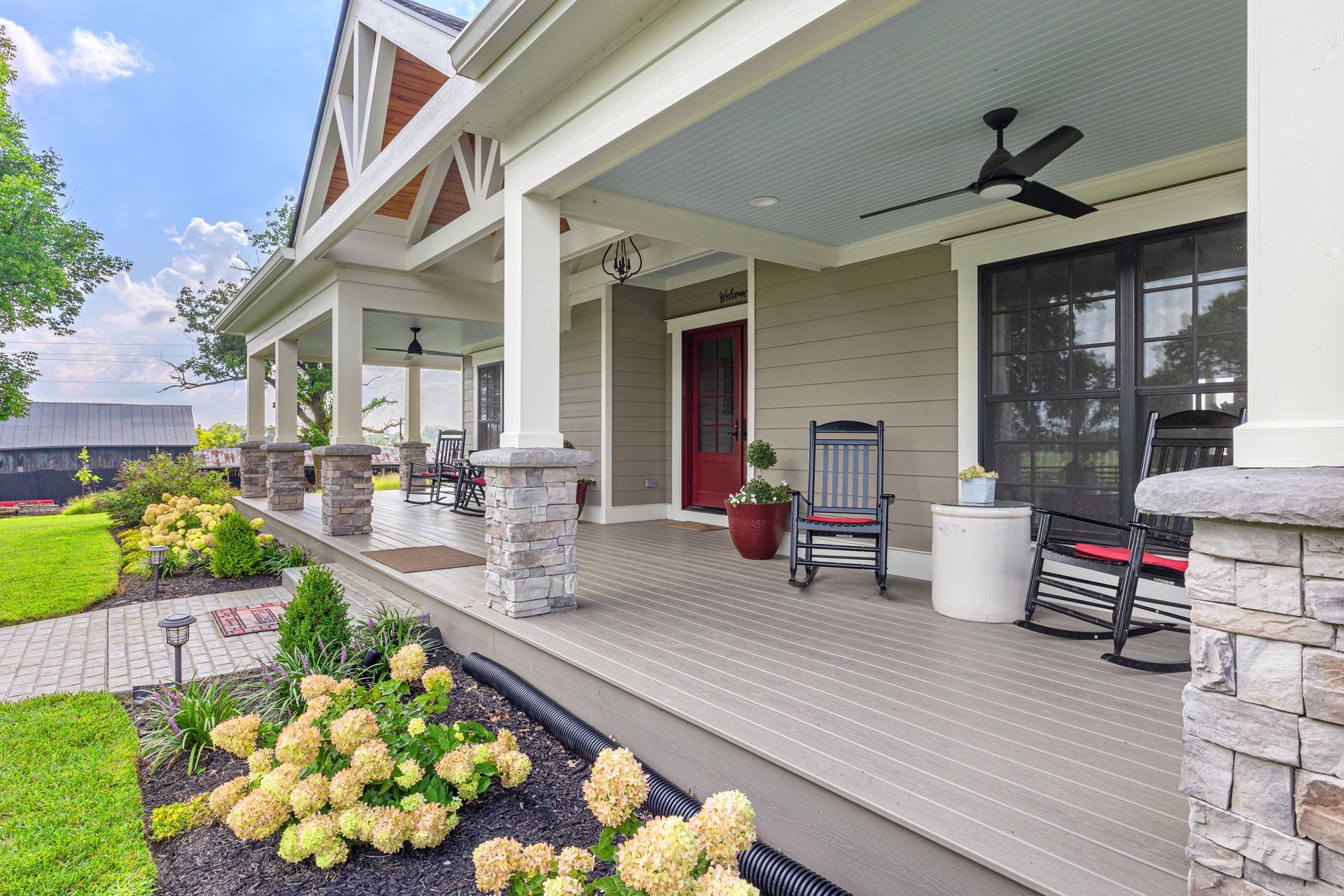 A long, covered front porch with gray siding, stone columns, and rocking chairs. Landscaping with flowers in front.