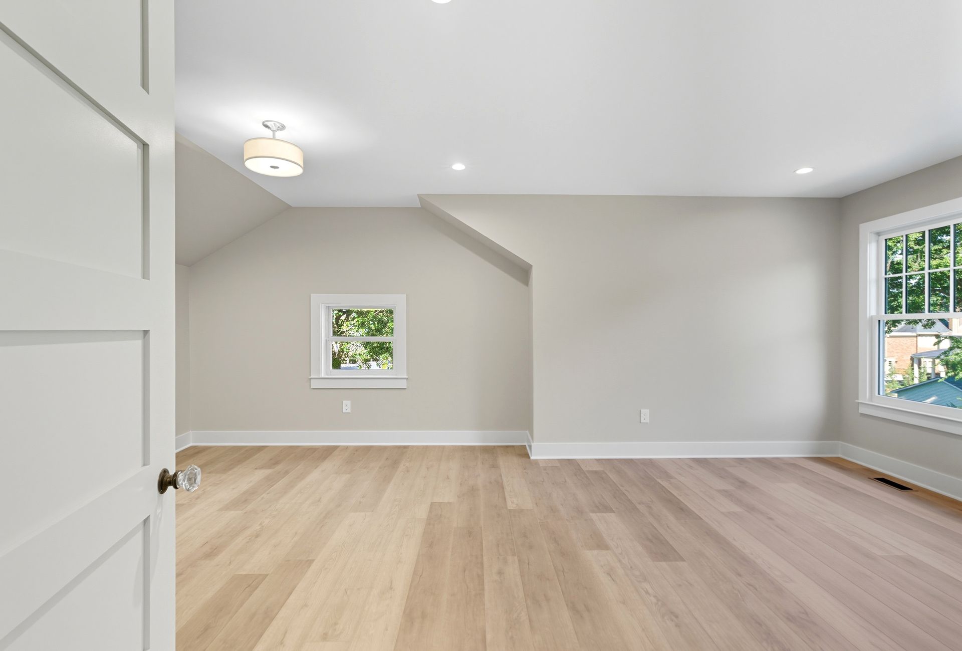 Empty, neutral-toned bedroom with hardwood floors, a small window, and a partially open white door.