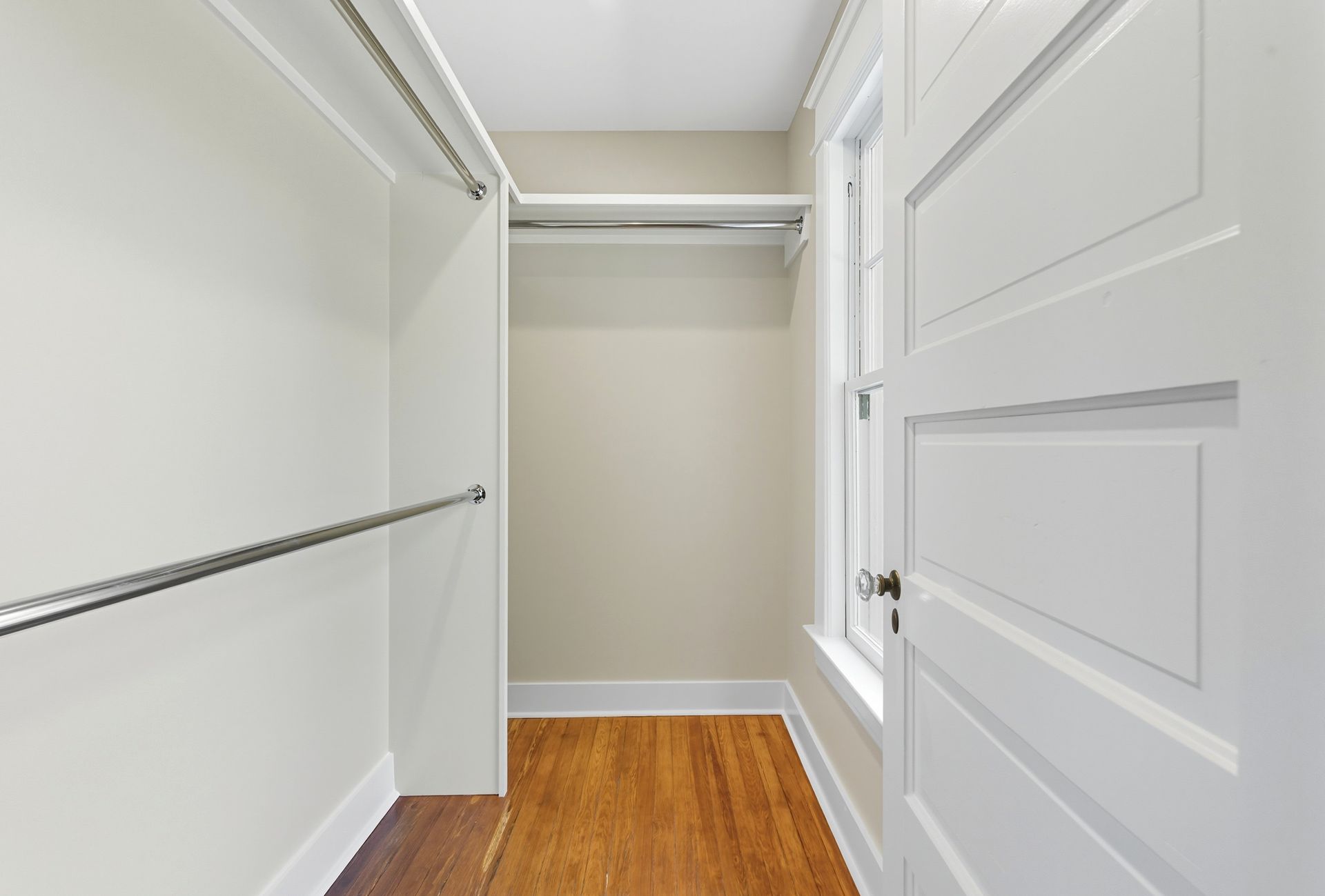 Empty walk-in closet with white walls, wooden floor, chrome rods, and white-framed window.