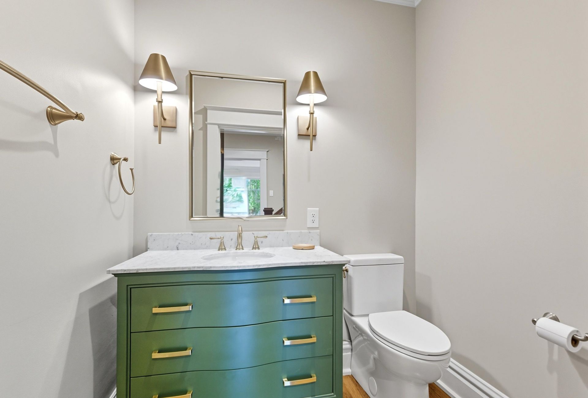 A green bathroom vanity with a white countertop and gold fixtures. There is a mirror, sconces, and a toilet.
