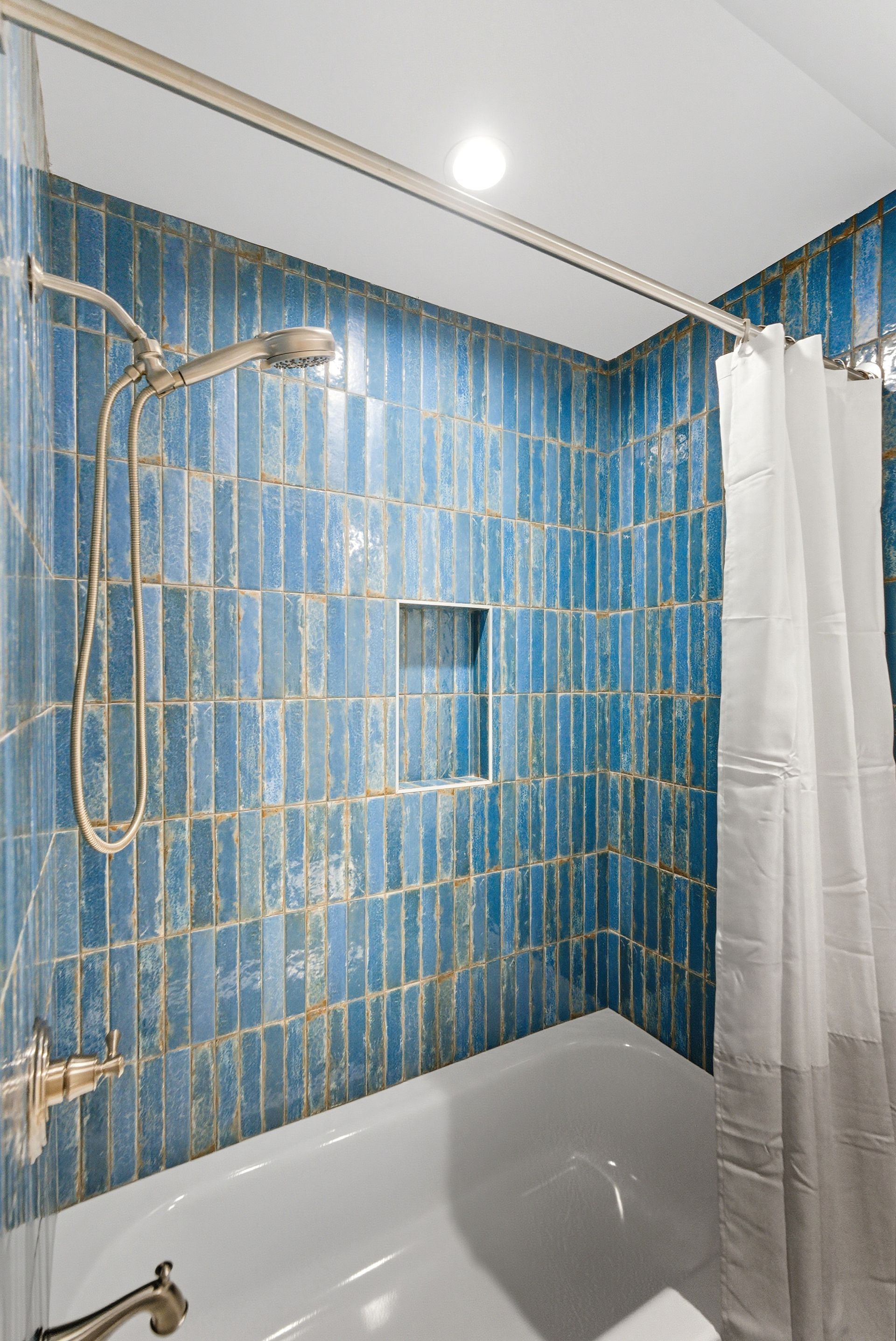 Blue tiled shower with a white bathtub and curtain.