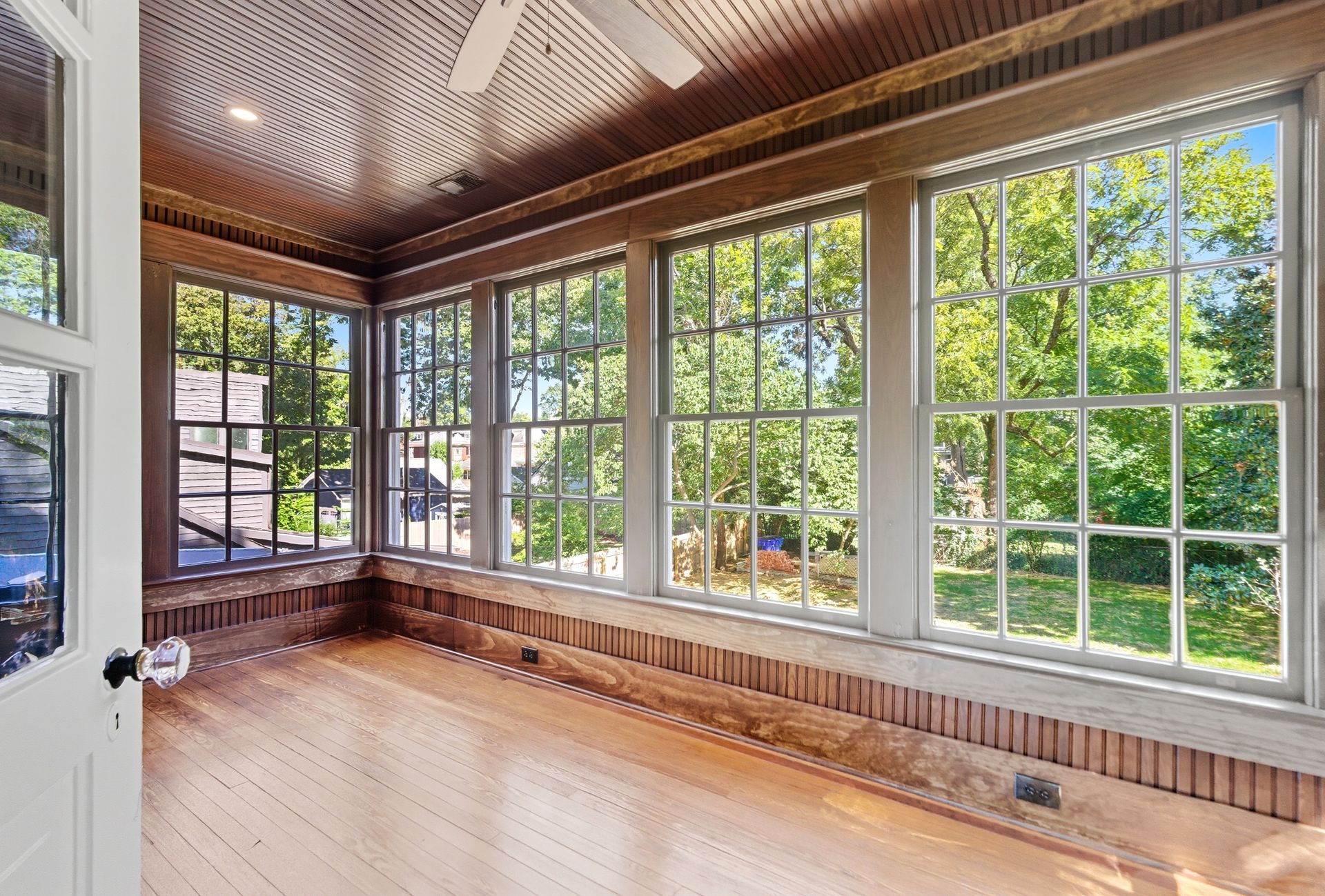Sunroom with large windows, wooden floor, and ceiling, overlooking a green yard.