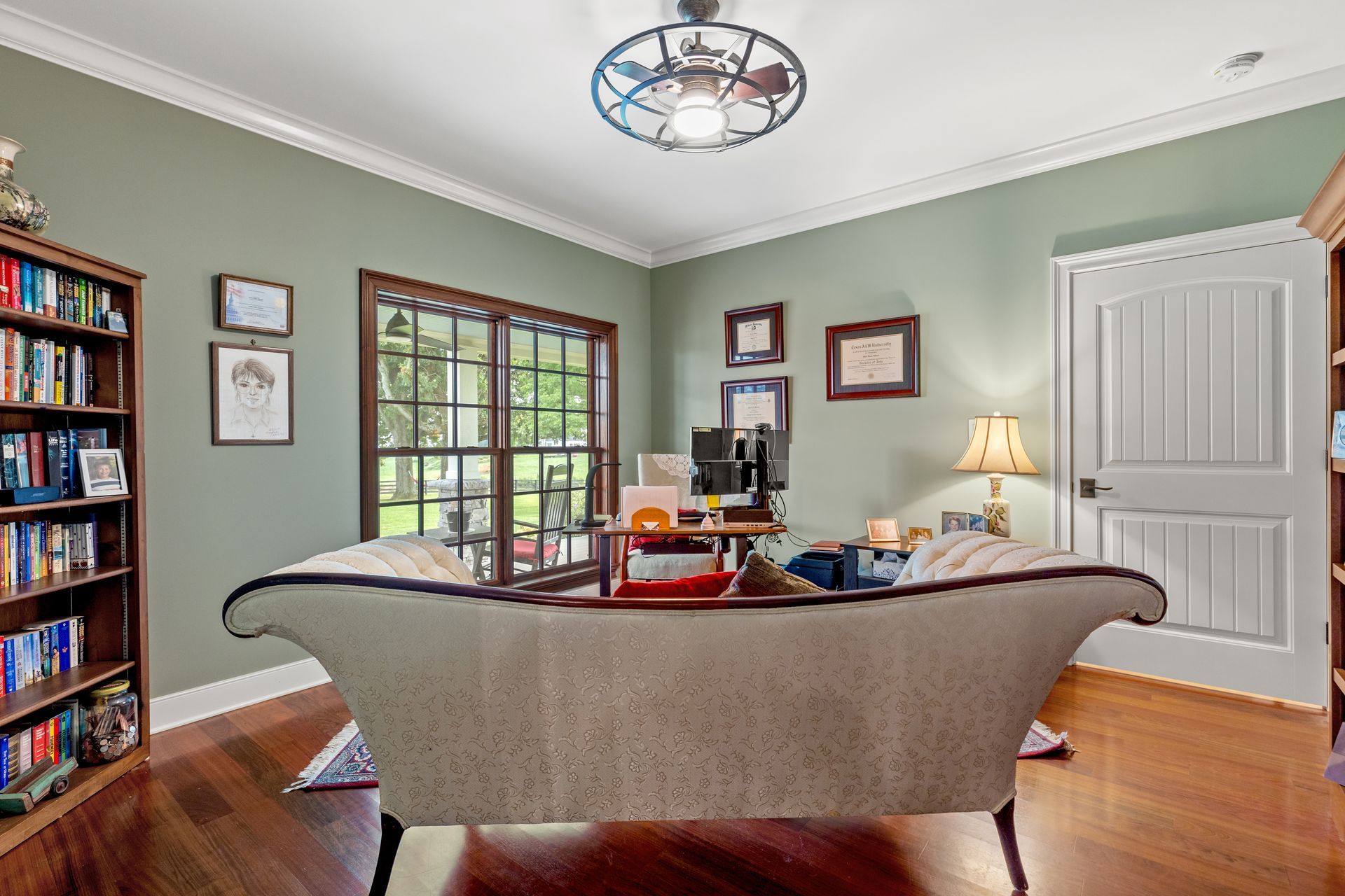 Office with sage green walls, hardwood floors, bookshelves, and a sofa. Desk by the window; door on the right.