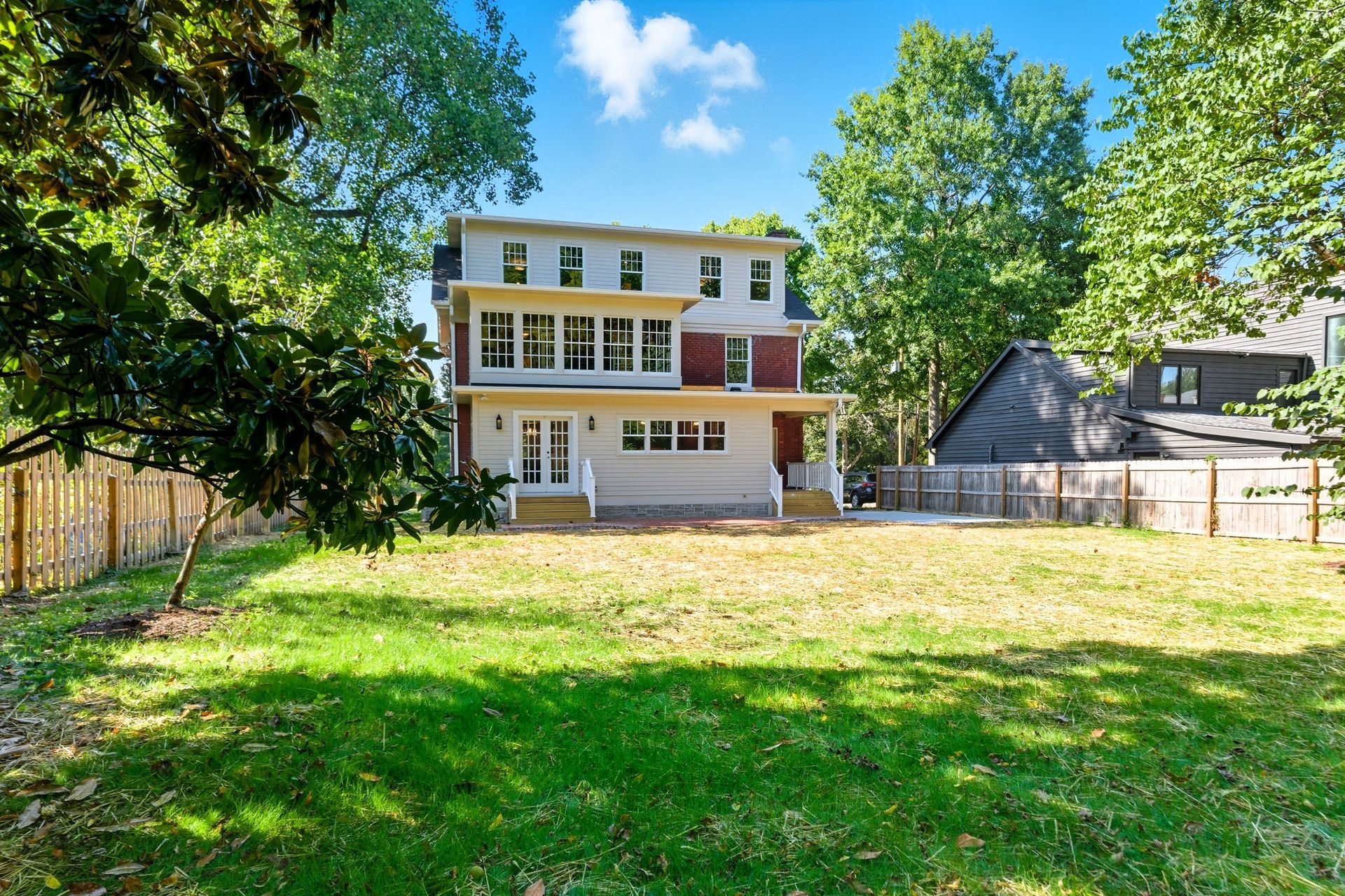 Three-story house with a large grassy yard surrounded by trees and a wooden fence. Sunny day.