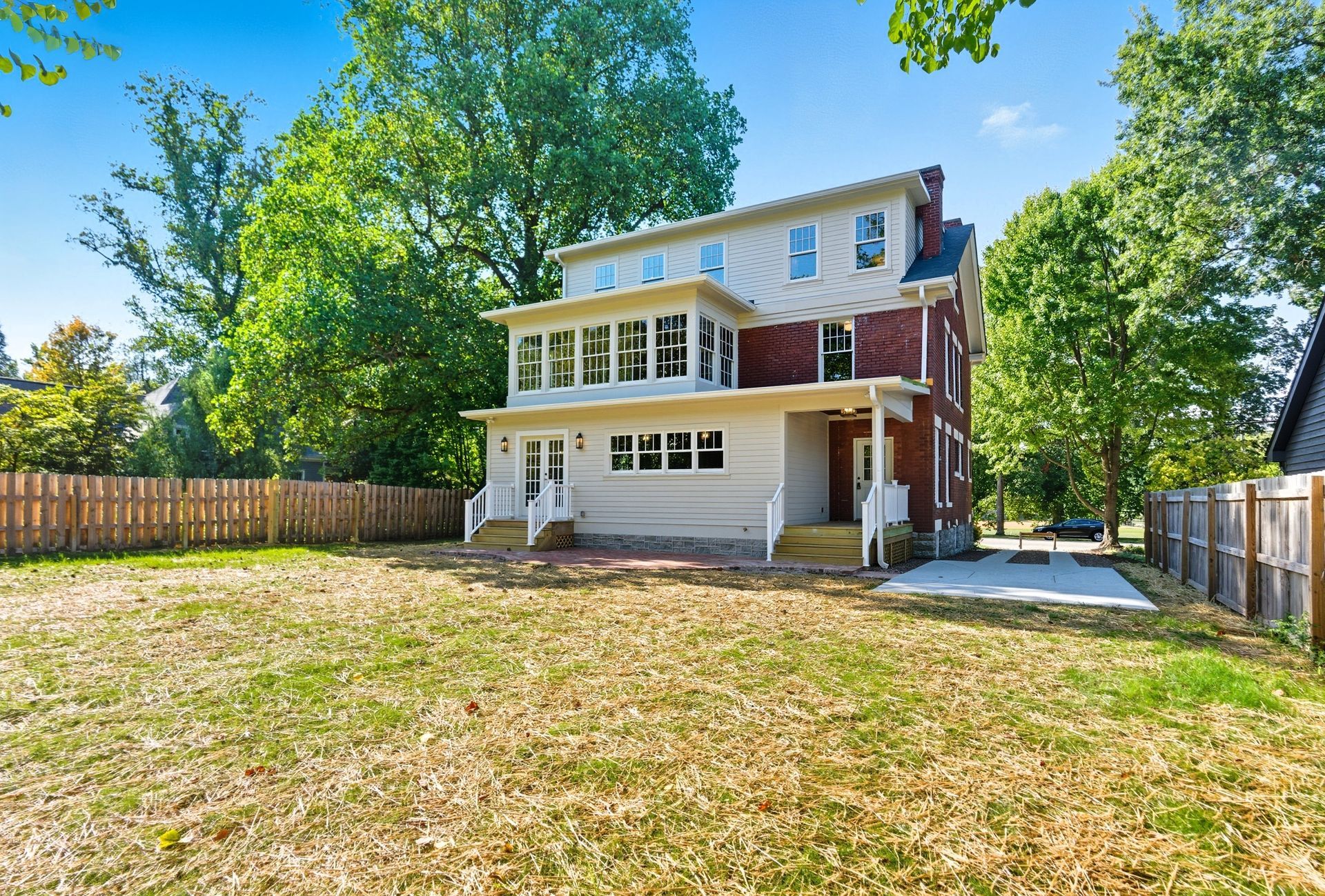 Back view of a two-story house with a large yard and wooden fence; trees and blue sky in the background.