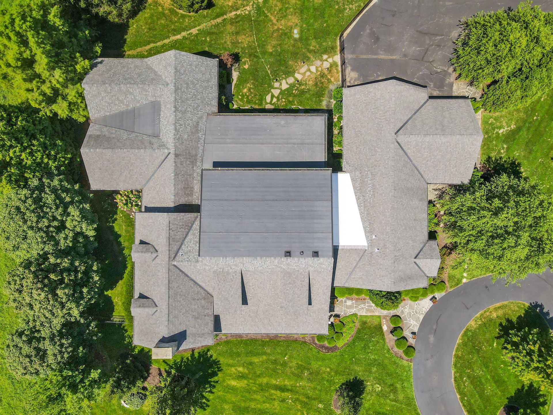 Aerial view of a house with a gray roof surrounded by green grass and trees. A curved driveway is visible.