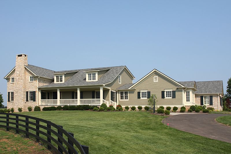 Large house with light brick, green siding, and a wraparound porch, on a green lawn with a black fence.