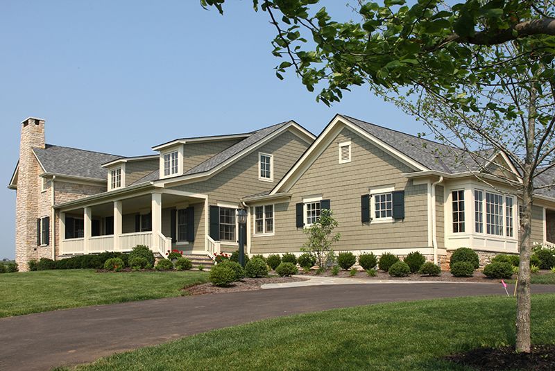 Large, green-sided house with stone chimney, wraparound porch, and black shutters on a sunny day.