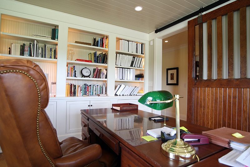 A home office with a wooden desk, leather chair, bookshelves, and a green lamp.