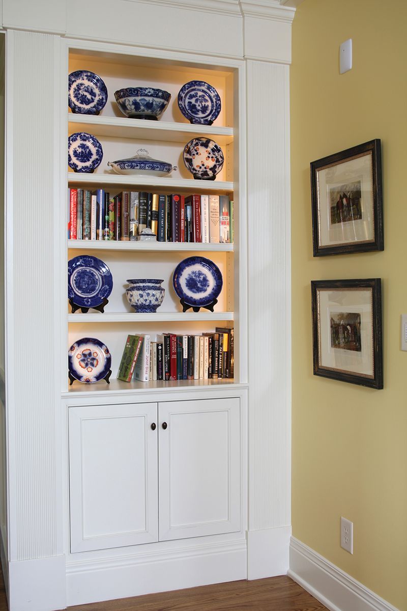 White built-in bookcase with blue and white plates and books. Yellow wall with two framed photos.