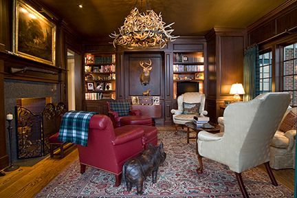 Cozy study: dark wood paneling, leather armchairs, deer head, antler chandelier, fireplace, and bookshelves.