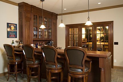 Elegant home bar with wooden cabinets, stools, and French doors; warm lighting.