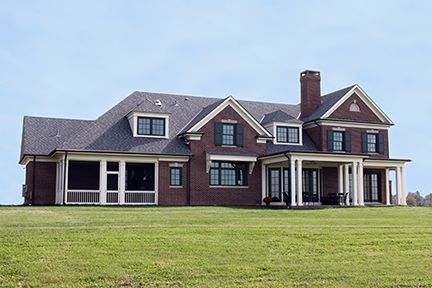 Large brick house with black shutters, porch, and chimney on a green lawn under a blue sky.