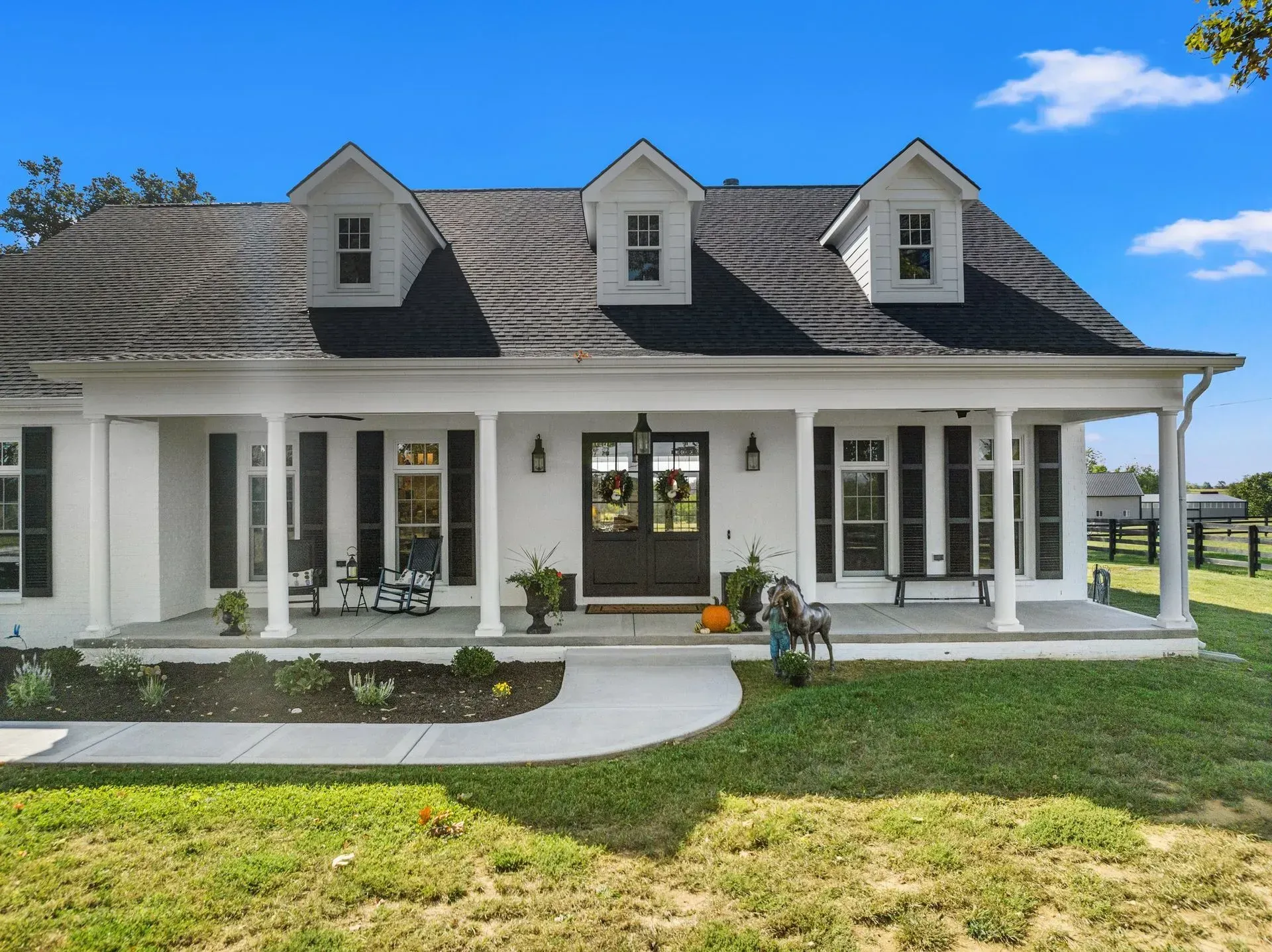 White farmhouse with black shutters, dormers, and a porch, set on a grassy lawn under a blue sky.