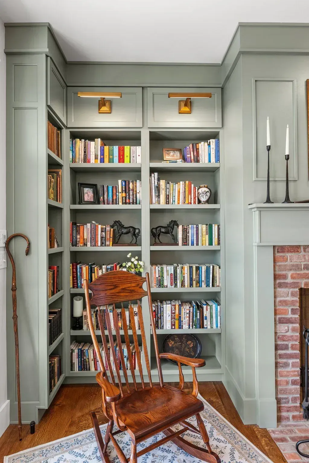 A cozy reading nook with green bookshelves, a wooden rocking chair, and a brick fireplace.