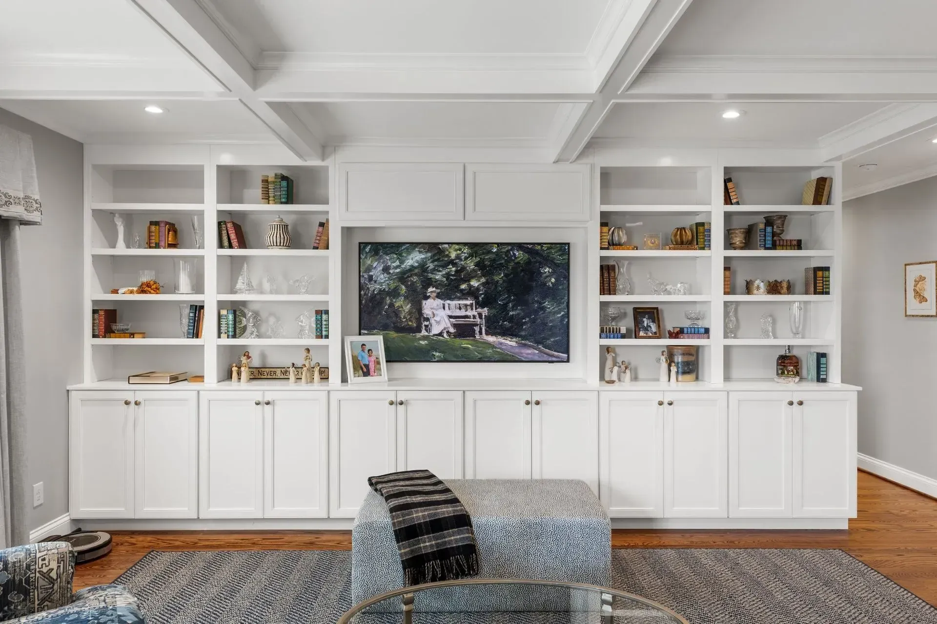 White built-in shelves and cabinets surround a TV in a gray-walled living room; a blue ottoman sits in front.