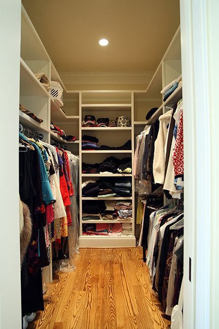 Narrow walk-in closet with white shelves, hanging clothes, and wooden floors.