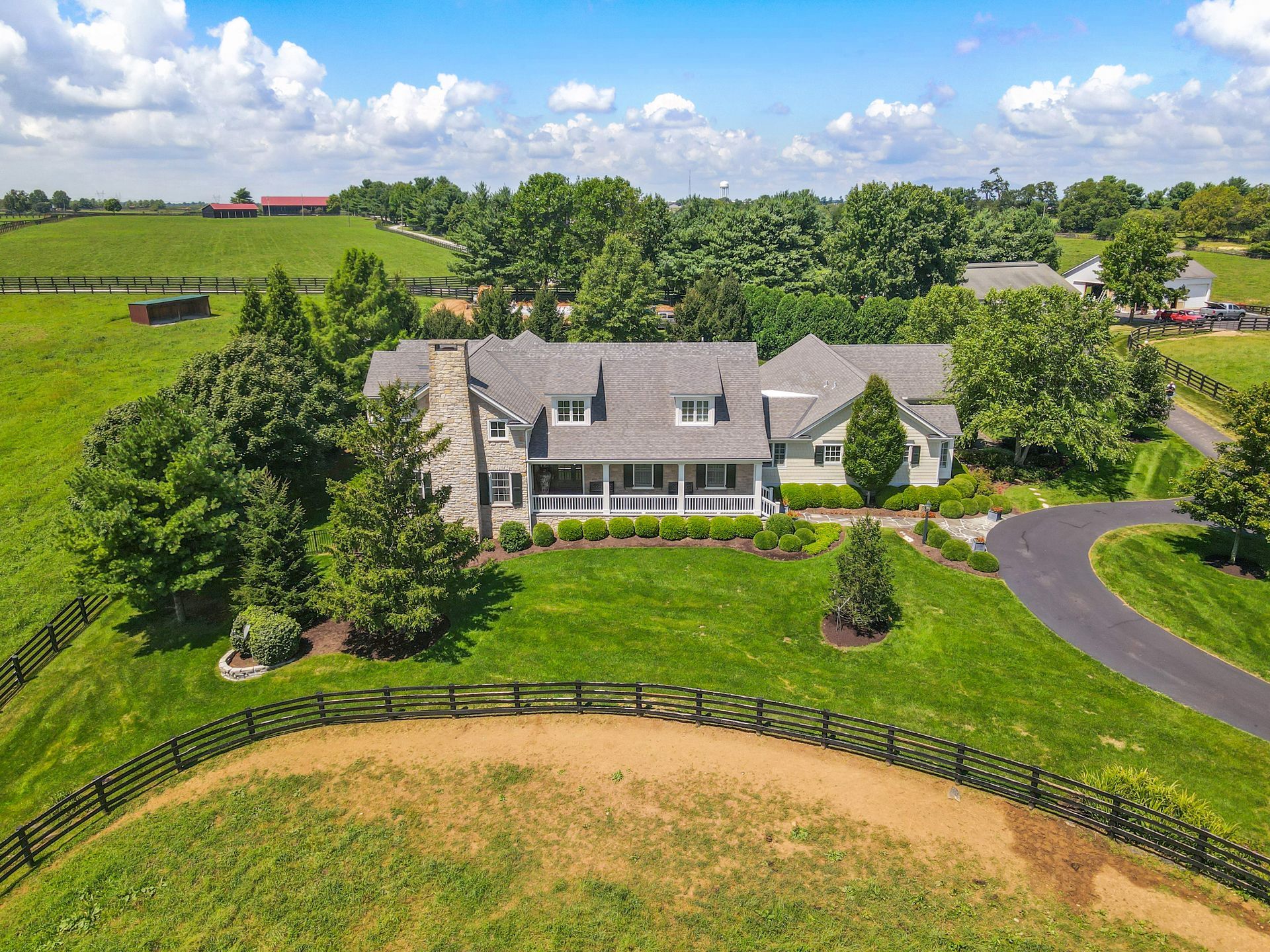 Sprawling house on a green farm surrounded by fields, trees, and black fencing under a blue sky.