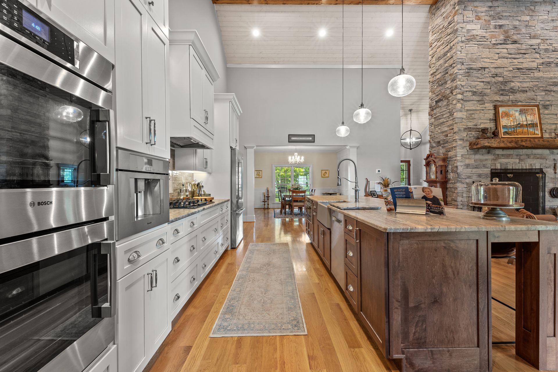 A modern kitchen with white cabinets, stainless steel appliances, a stone fireplace, and a long island.